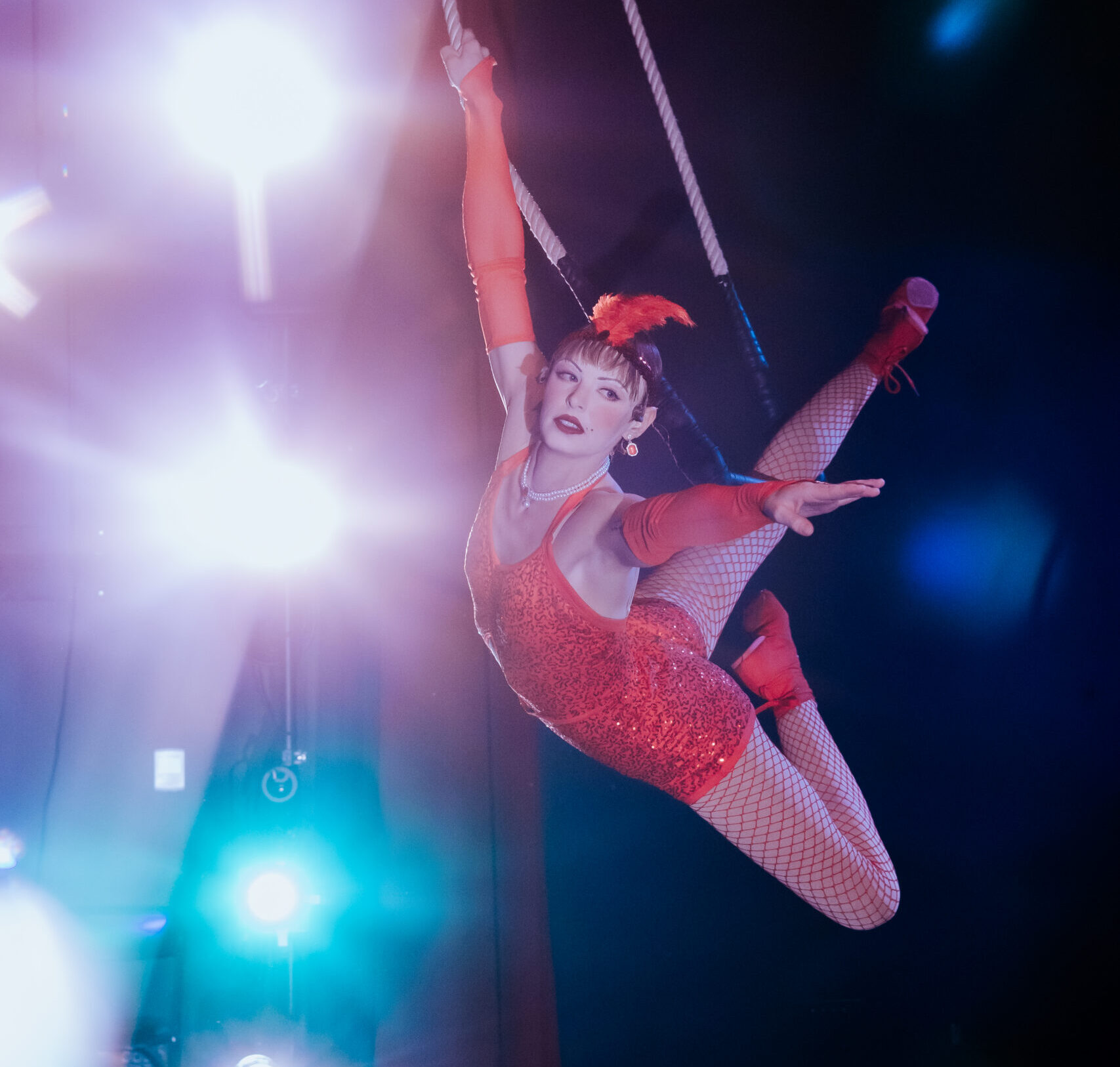 adult performance company student on a trapeze flying through the air smiling at the camera. She is in a red sparkle dress that looks like a flapper.