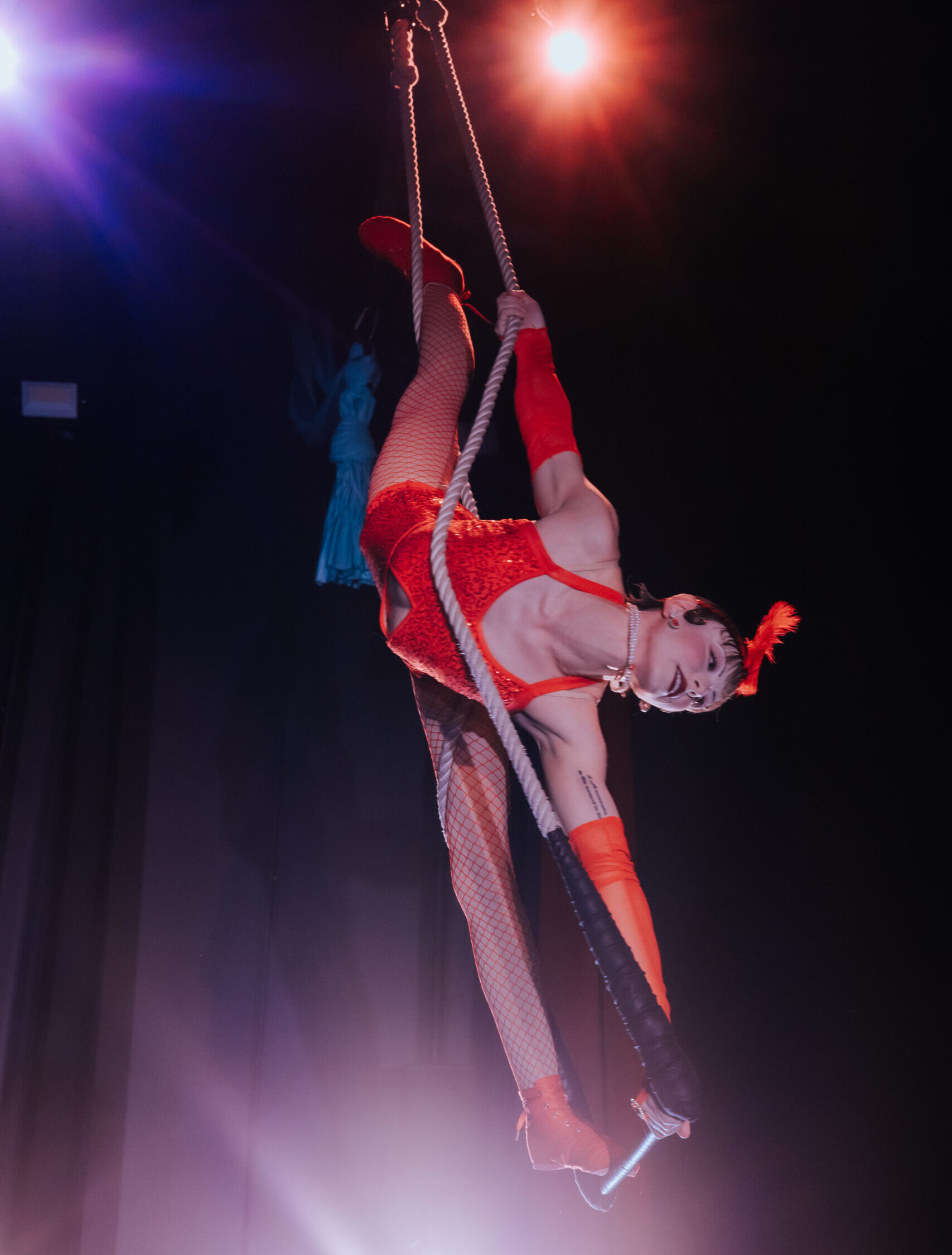 A trapeze artist performs at cleo parker robinson dance studio. She is wearing a red outfit and is in a split pose.