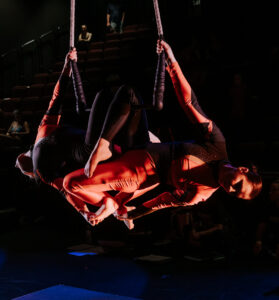 2 aerialists perform a duo trapeze act. They are hanging on the trapeze bar by 1 kneee each.