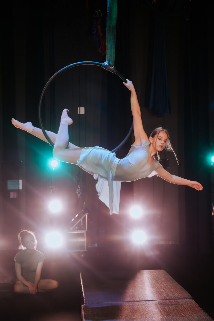 A teen aerial dancer showcases a lyra act at cleo parker robinson dance studio. She is wearing a light blue costume and had an arm outstretched.