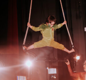 A youth student performs a trapeze act at aerial cirque over denver student showcase. He is 4 years old and performing a giant X shape on trapeze in a green costume.